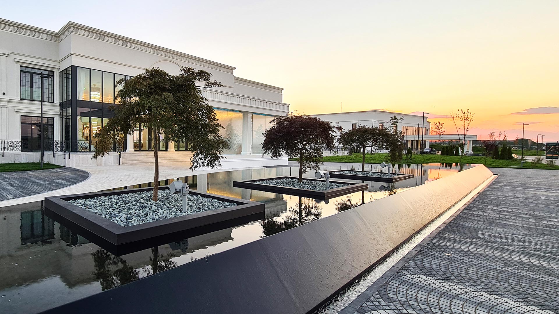 Grup de arțari decorativi plantați în insule rotunde, reflectați în apă la apus, în cadrul proiectului Ramatuelle.