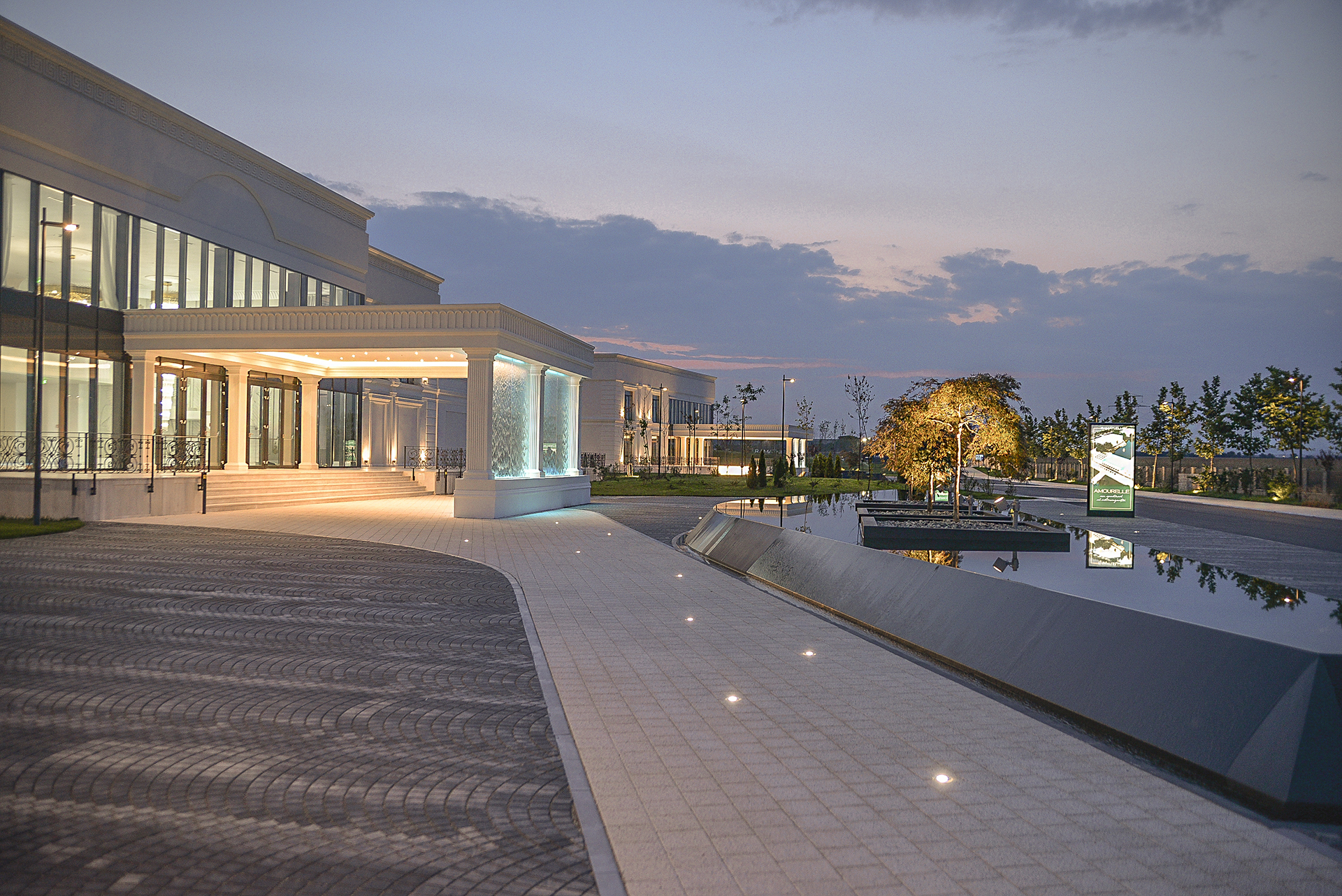 Intrare principală iluminată a pavilionului de evenimente Ramatuelle, înconjurată de apă și vegetație ornamentală